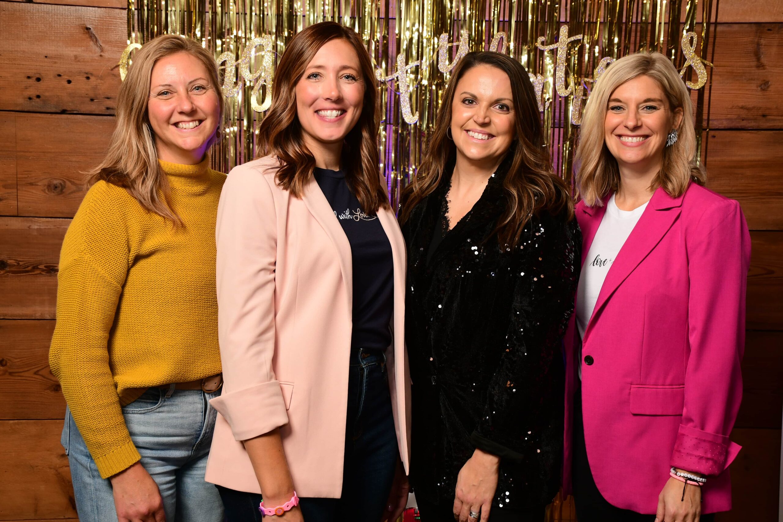 Four women smile at the camera in front of a shimmering background